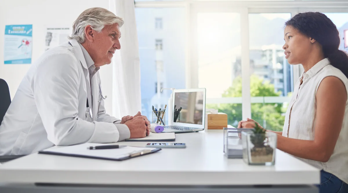 A doctor and a patient sitting across a desk in a bright consulting room, talking