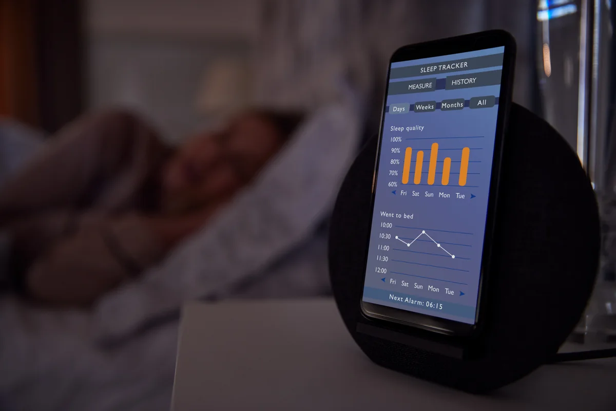A phone on a nightstand showing five nights of sleep quality and bedtimes while someone sleeps behind it
