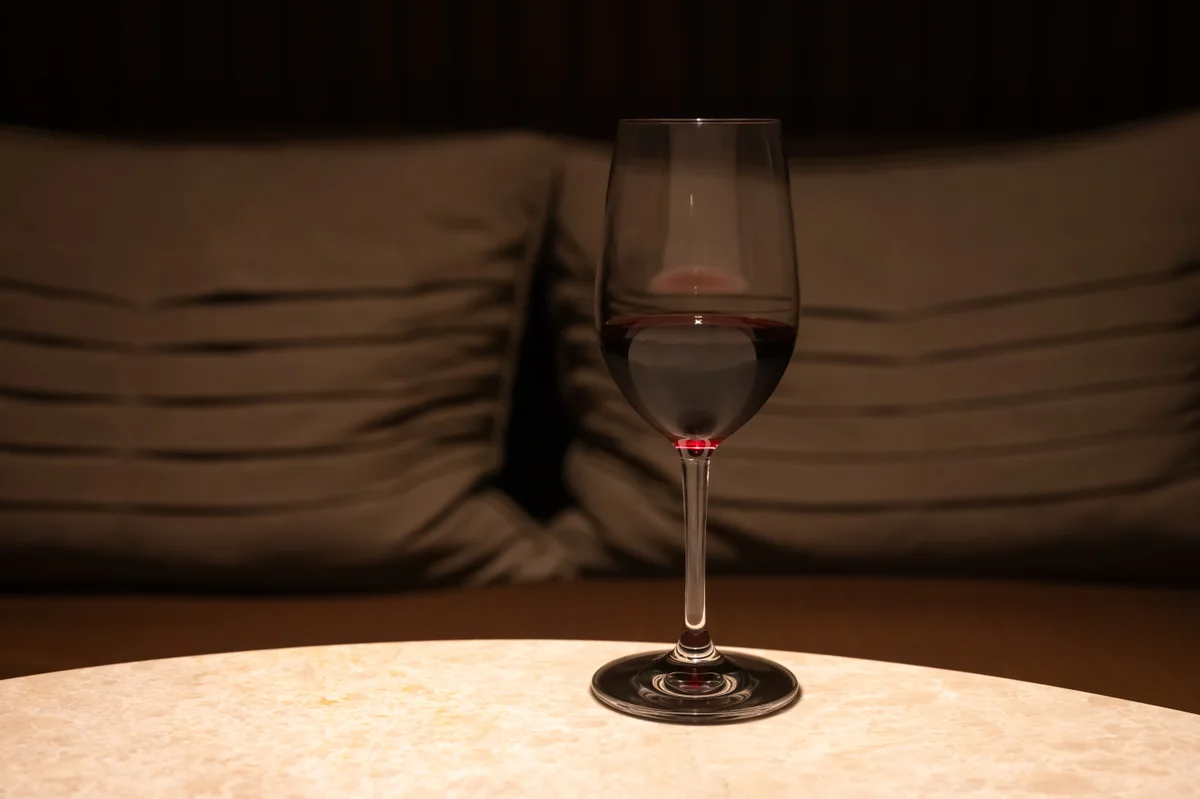 A single glass of red wine on a table in low light, with cushions behind it