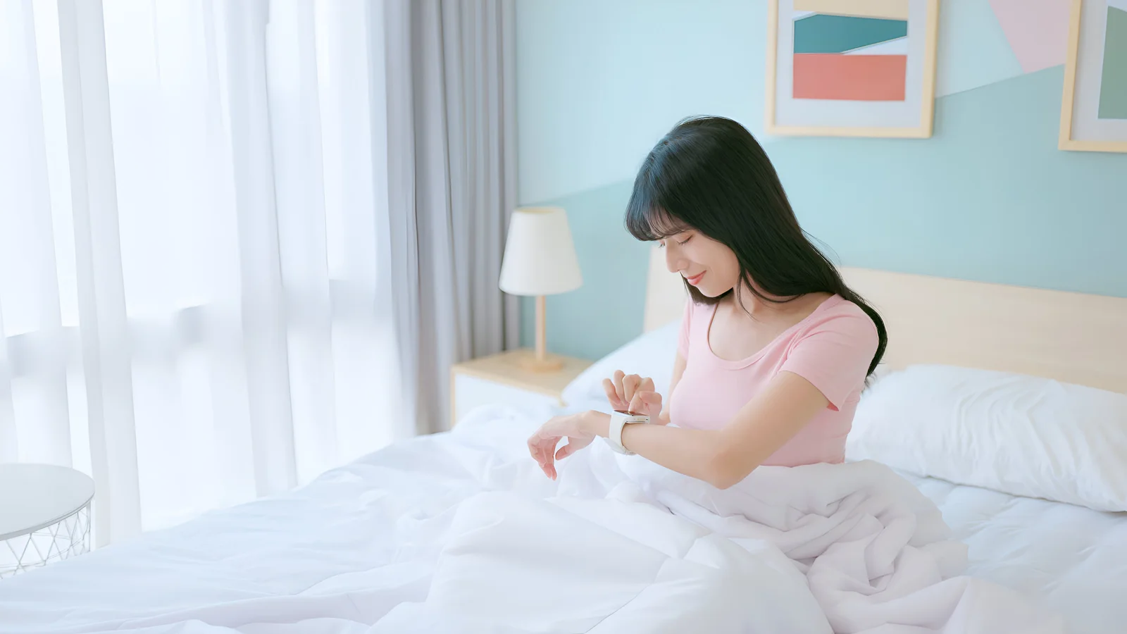 A woman sitting up in bed in morning light, looking at the smartwatch on her wrist