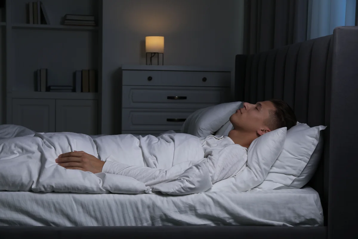 A man asleep on his side in a dim bedroom with a lamp on the nightstand