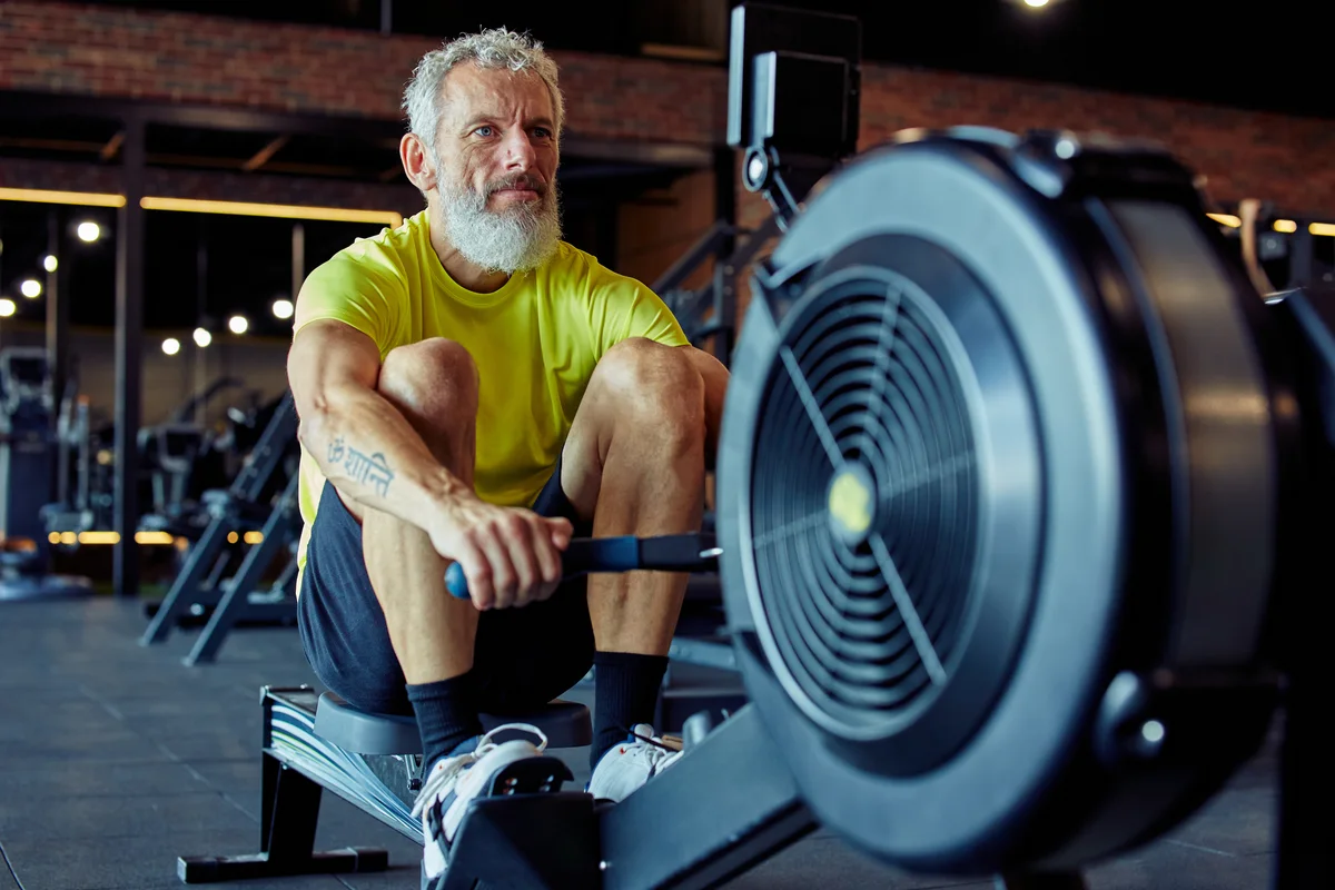 Man with a white beard resting forward on an indoor rowing machine, the flywheel filling the foreground