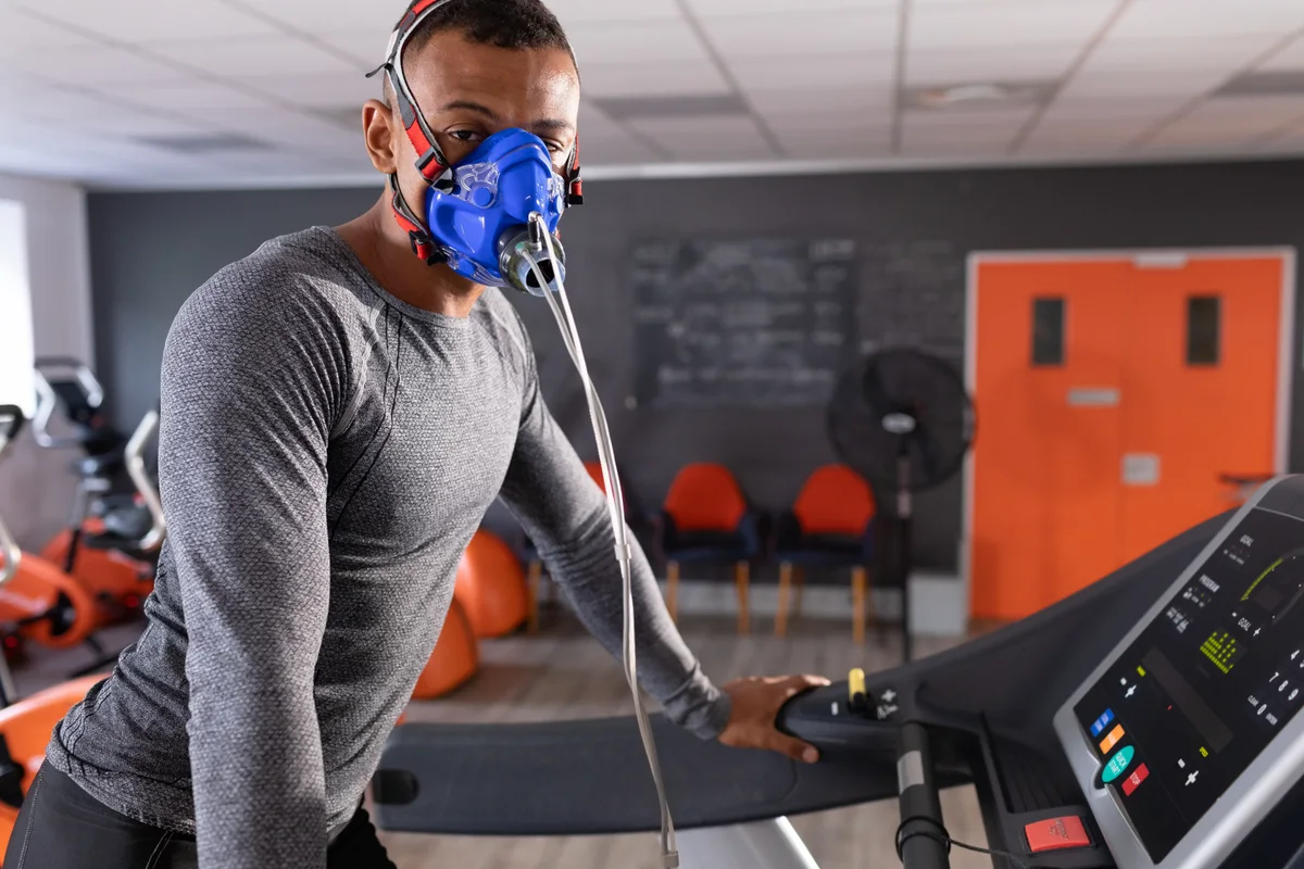 A man leans on a treadmill console breathing through a blue gas mask, its sampling line running to the floor